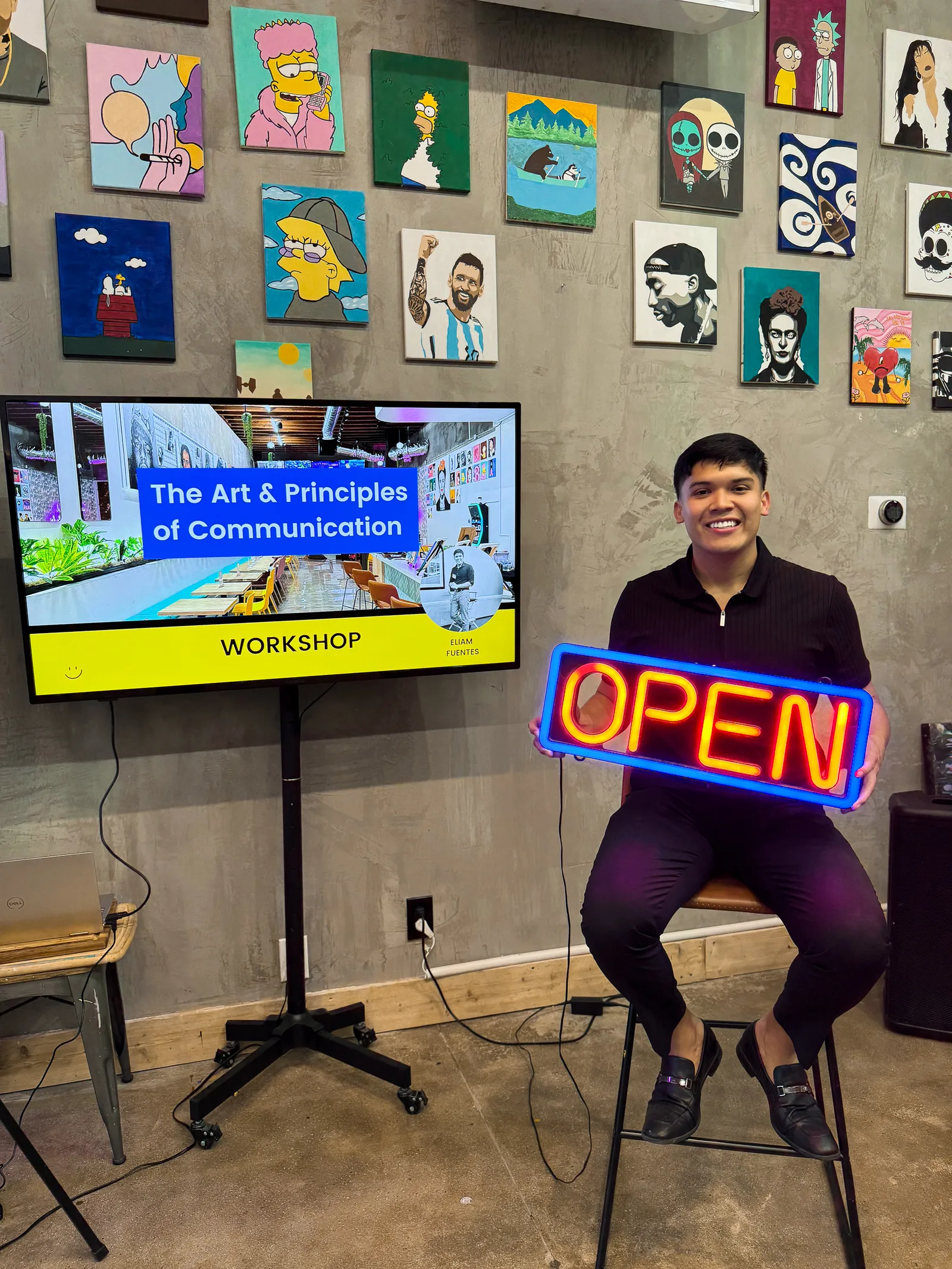 Eliam holding an OPEN sign beside his communication workshop screen